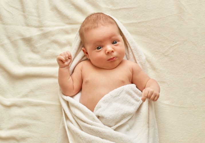 Newborn baby wearing towel after bath, cute tiny child lying on bed on white blanket, looking and studying outward things. Childhood and baby care.